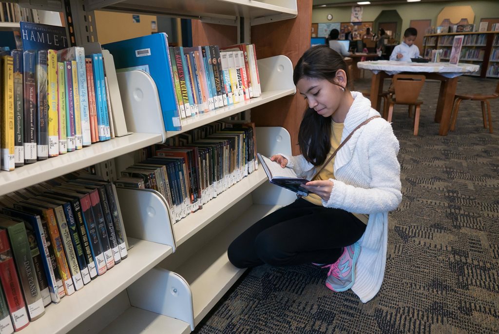 A young girl reading a book off a library shelf