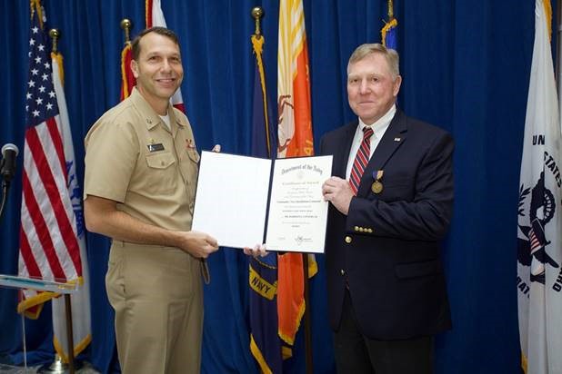 Captain Brian Weiss presents award to Harrison Conyers III in front of flags