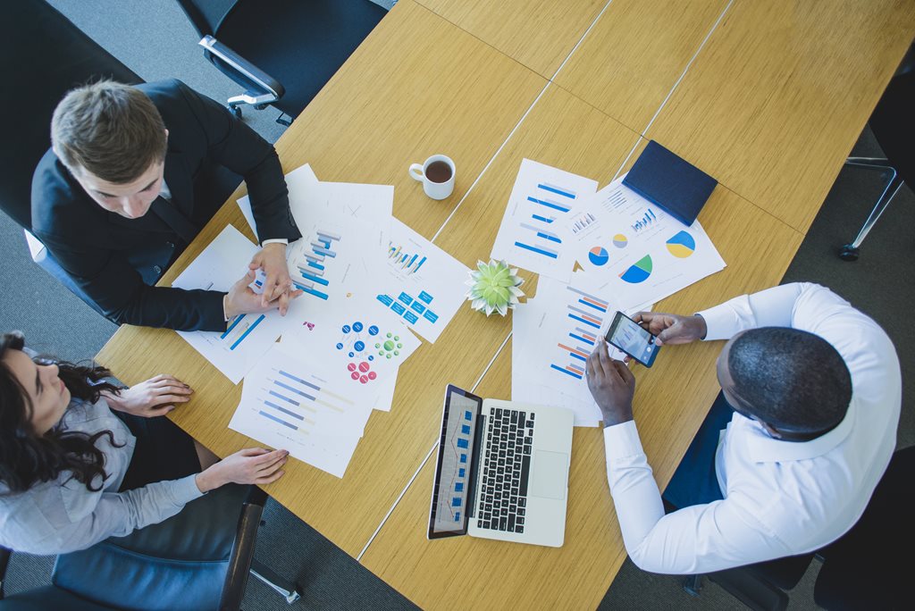 overhead photo of three business people sitting at conference table with computers and paperwork